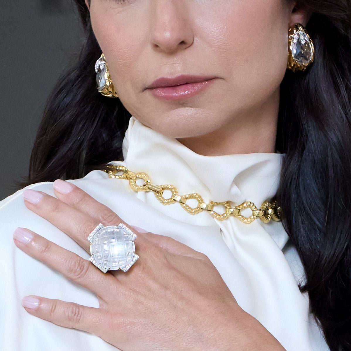 Woman wearing a large crystal David Webb ring and gold jewelry against a neutral background