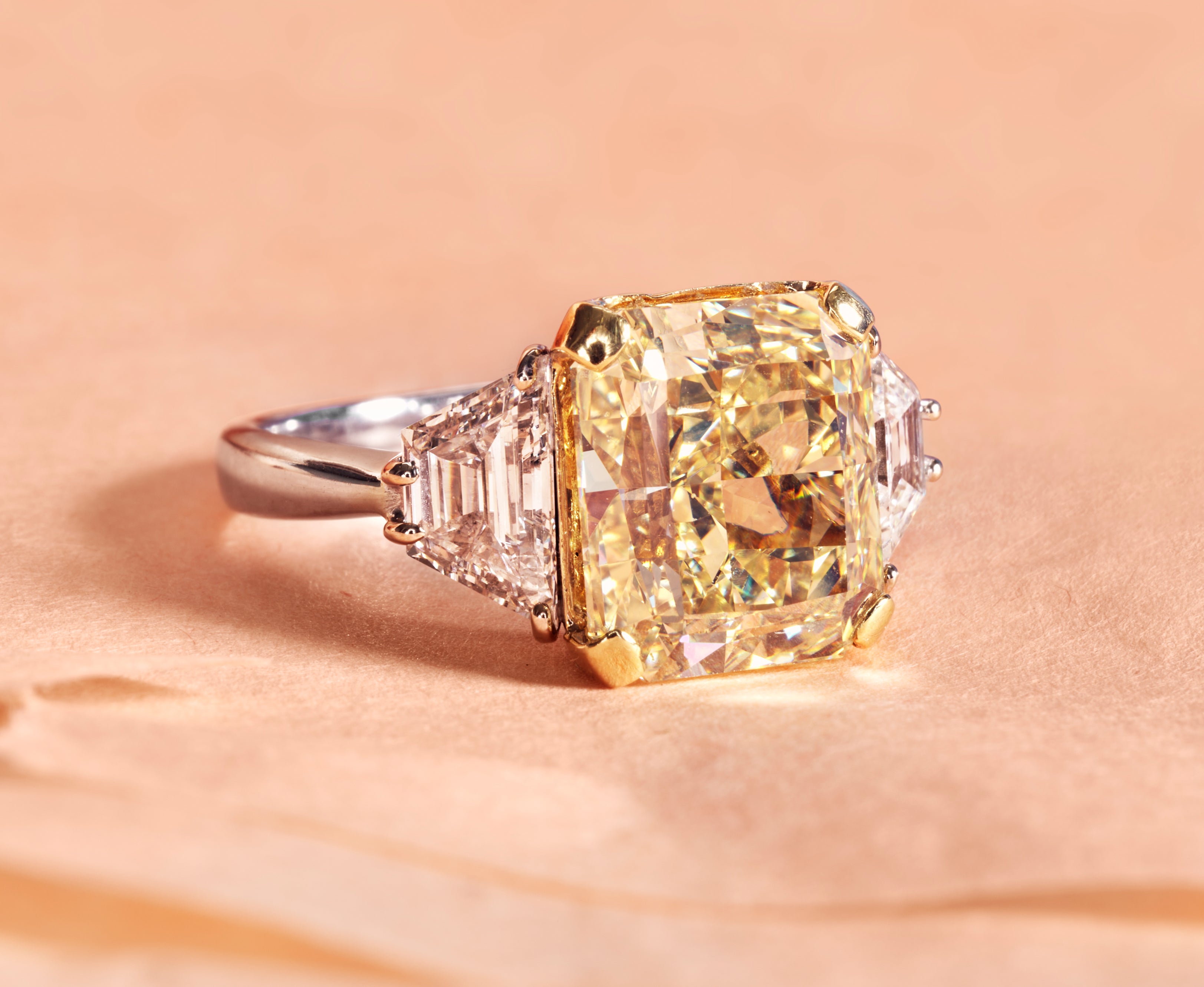 A diamond ring featuring a large cushion-cut yellow diamond center stone with baguette side stones and a silver-colored band, resting on a textured peach-beige surface.